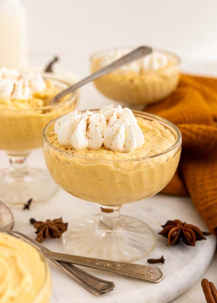 A glass dessert dish filled with creamy pumpkin mousse, topped with whipped cream and a sprinkle of cinnamon. Other mousse dishes and spices are in the background.
