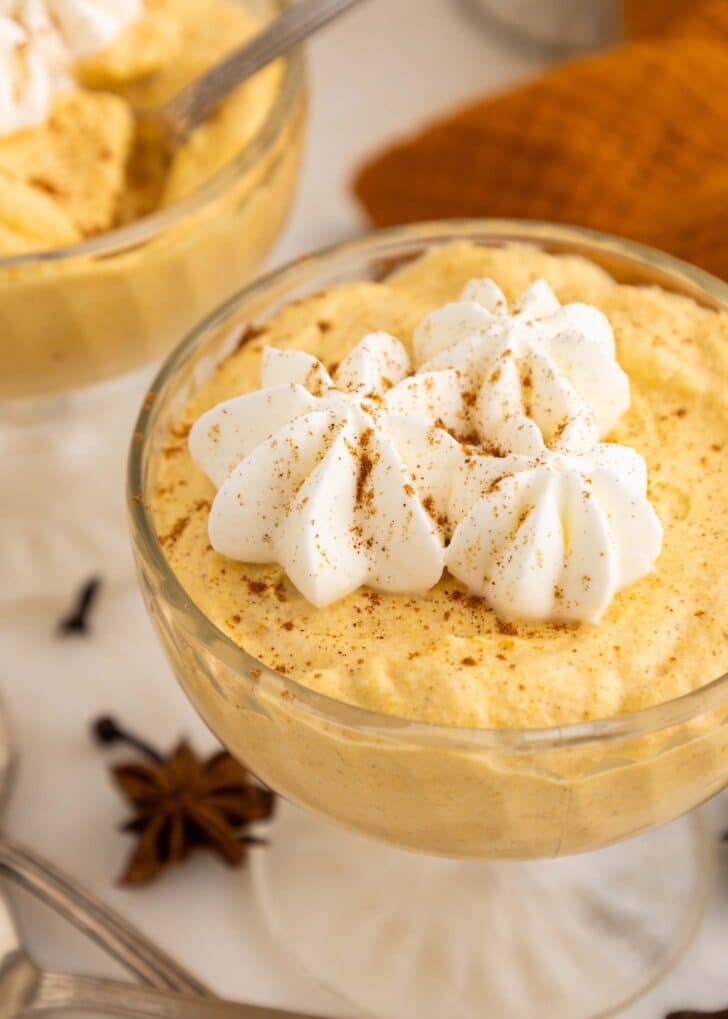 A glass dish filled with creamy pumpkin mousse, topped with swirls of whipped cream and a sprinkle of cinnamon, with star anise and a brown napkin in the background.